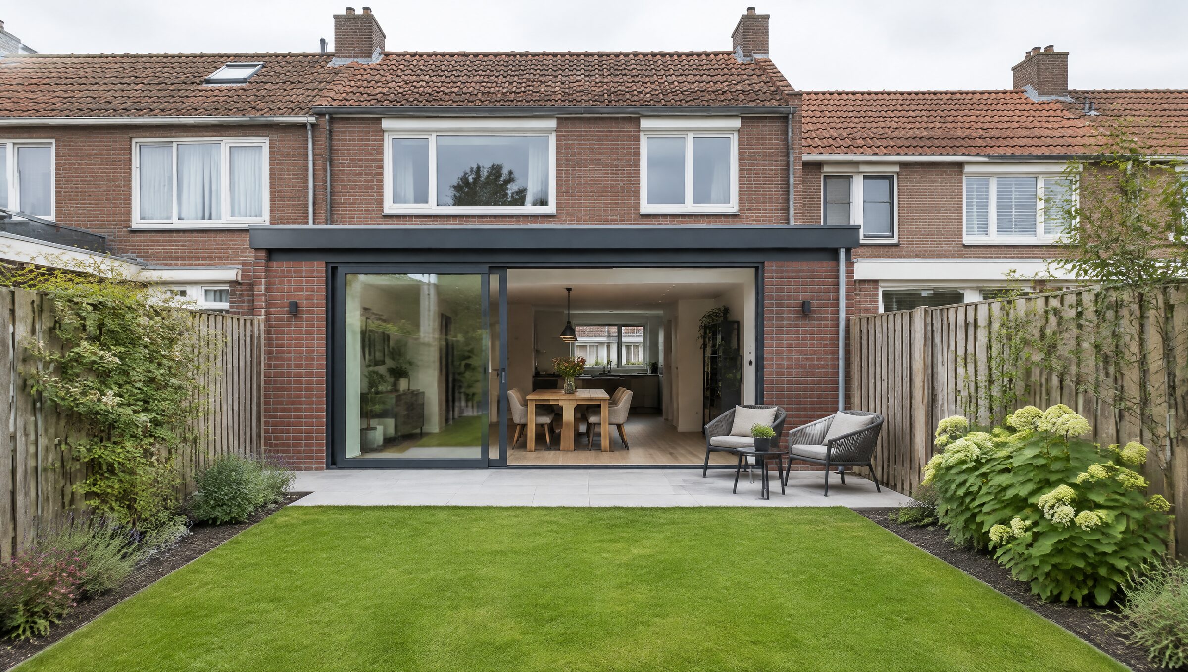 De achterkant van een Nederlands rijtjeshuis van rode baksteen met een nieuwe uitbouw: plat dak, grote glazen schuifpui en een eettafel binnen, gezien vanaf het gazon op een heldere dag.